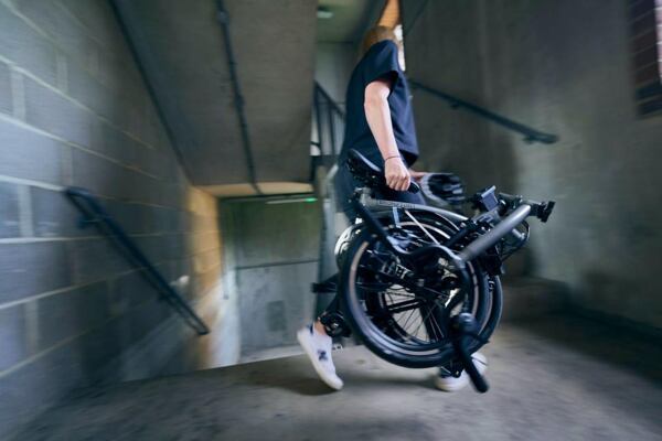 Žena nese za sedlo složené skládací kolo Brompton do schodů